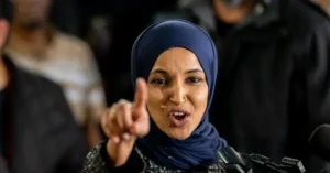 A woman wearing a hijab is speaking into a microphone, pointing forward with her right hand.
