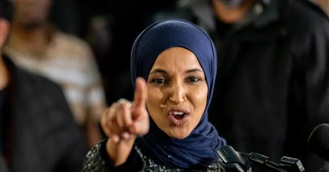 A woman wearing a hijab is speaking into a microphone, pointing forward with her right hand.