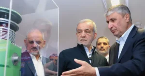The photo shows a group of men in a formal setting, possibly at a technological or industrial event, with one man gesturing towards a display.