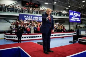 The image shows President Donald J. Trump at a rally in Alaska, where he is pointing towards the crowd, with a sign in the background reading "MAKE AMERICA GREAT AGAIN."