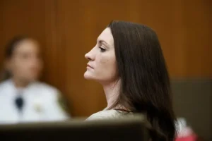 The image shows a woman with long dark hair seated in what appears to be a courtroom setting.