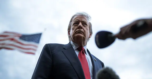 The image shows a man in a suit and red tie standing outdoors with an American flag in the background, suggesting a political or public event.