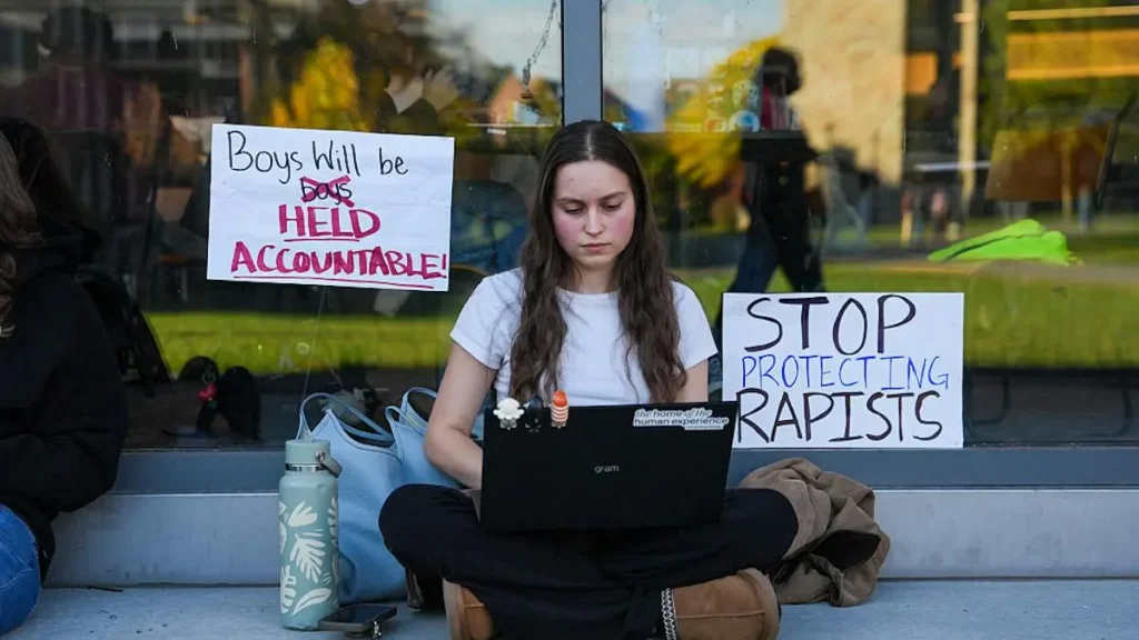 A person sits on the ground with a laptop, surrounded by protest signs advocating for accountability and stopping the protection of rapists.