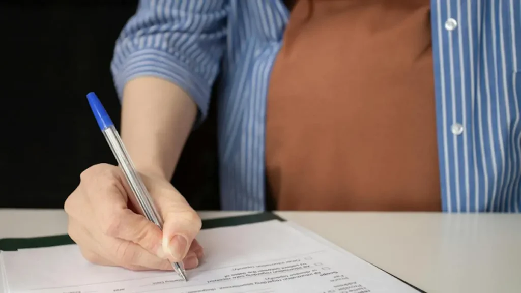 A person is signing a document with a blue pen at a desk.