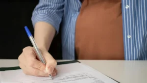 A person is signing a document with a blue pen at a desk.