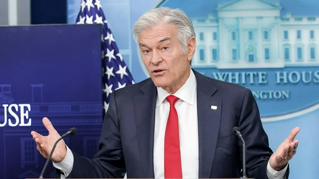 A man in a suit and red tie is speaking at a podium in front of a backdrop featuring the White House and an American flag.