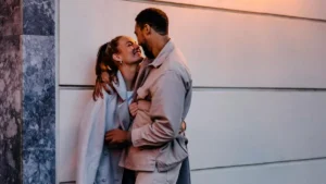 A couple shares a tender moment, embracing and smiling against a plain wall.