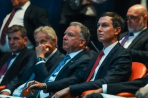 The image shows a group of men in formal attire seated in an audience, attentively watching a presentation or event.
