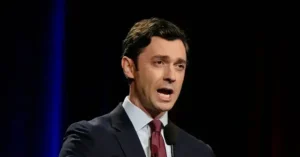 The image shows a man in a suit and tie speaking into a microphone, likely during a formal or professional event.