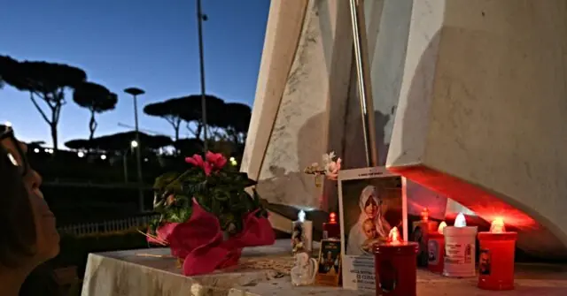 A memorial setup with candles, flowers, and a photograph is placed under a structure, with a person observing it in the foreground.