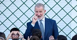 A man in a suit is speaking to a group of children in front of a green lattice fence.