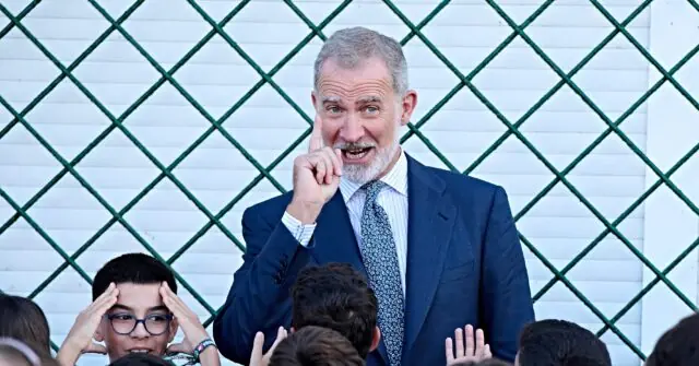 A man in a suit is speaking to a group of children in front of a green lattice fence.