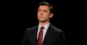The image shows a man in a suit and tie speaking into a microphone against a dark background.