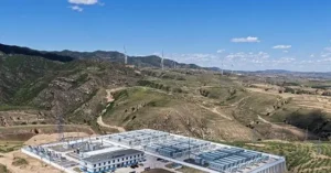 The image shows a large-scale renewable energy facility with wind turbines in the background, set against a scenic mountainous landscape under a clear blue sky.