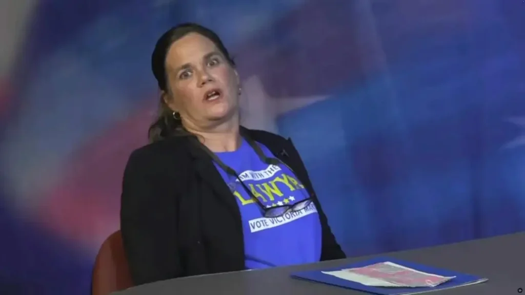 A woman wearing a blue shirt with political campaign text is seated at a table, appearing to speak or react to something.