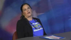 A woman wearing a blue shirt with political campaign text is seated at a table, appearing to speak or react to something.