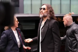 A man in a suit and sunglasses is seen interacting with another individual in a pinstriped suit, with a black vehicle and another person in the background.