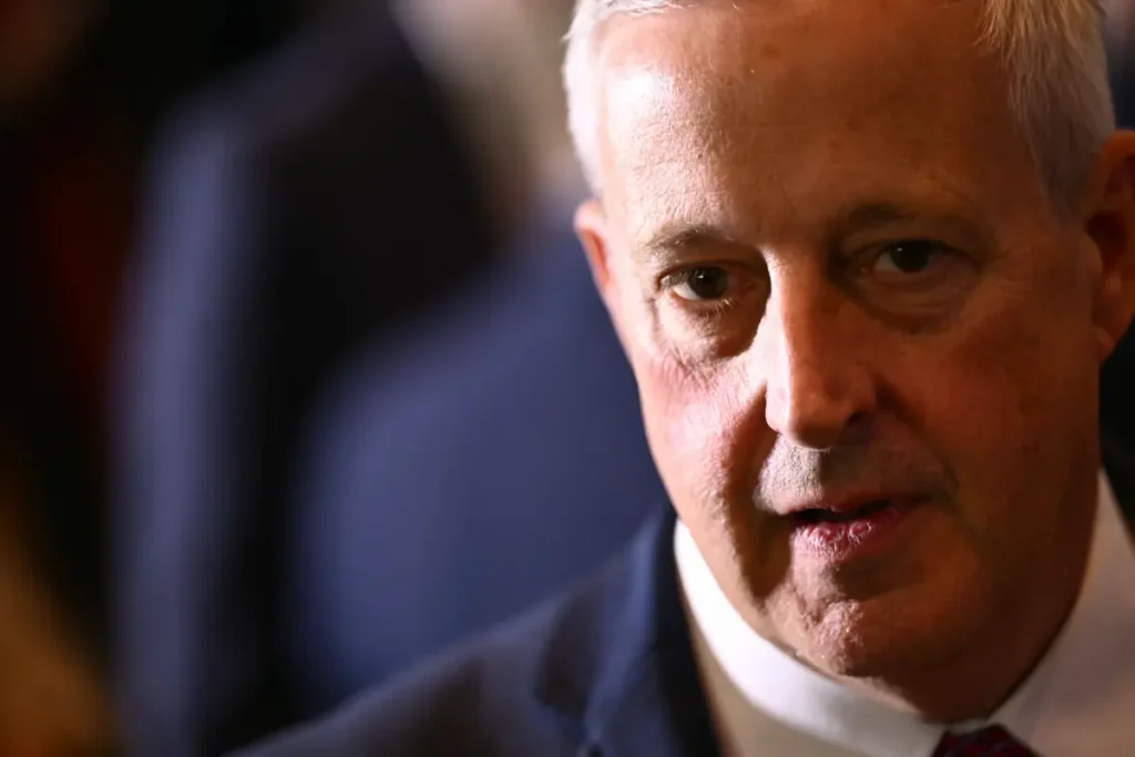 The image shows a close-up of a man in a suit, appearing to be speaking or listening intently.