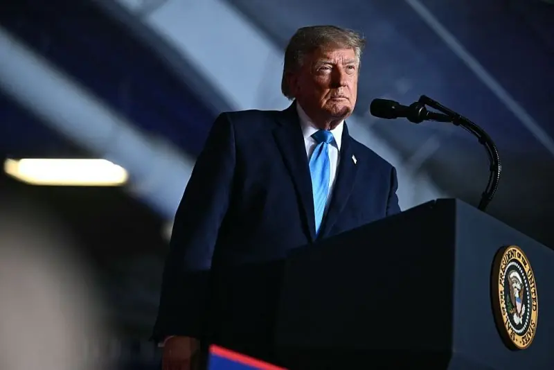 The image shows a man in a suit standing at a podium with the presidential seal, delivering a speech.