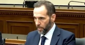 The image shows a man in a suit and tie, seated in what appears to be a formal setting, possibly a courtroom or a legislative chamber.