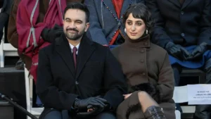 A man and a woman are seated together at an outdoor event, both dressed in winter attire, with the man wearing a suit and the woman in a brown coat.