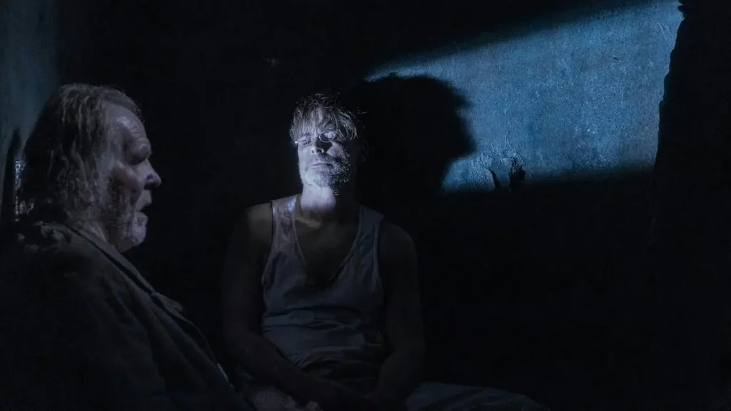 The image depicts two individuals in a dimly lit, possibly underground setting, engaged in a serious conversation.