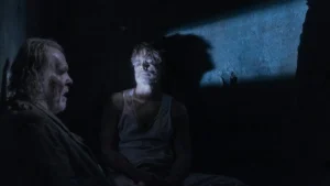 The image depicts two individuals in a dimly lit, possibly underground setting, engaged in a serious conversation.