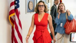 A woman in a red dress walks confidently in a formal setting with an American flag and other officials in the background.