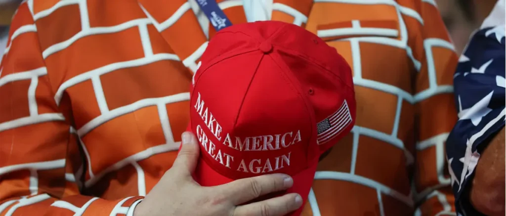 A person holds a red "Make America Great Again" hat in front of a brick-patterned shirt and an American flag.