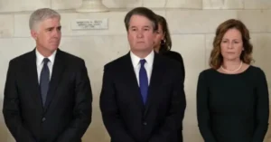 The image shows three individuals standing in formal attire, likely at a formal or official event.