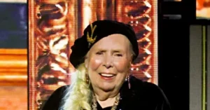 The image shows a person with long blonde hair styled in a braid, wearing a black outfit and a decorative headpiece, smiling in front of a backdrop with a golden design.