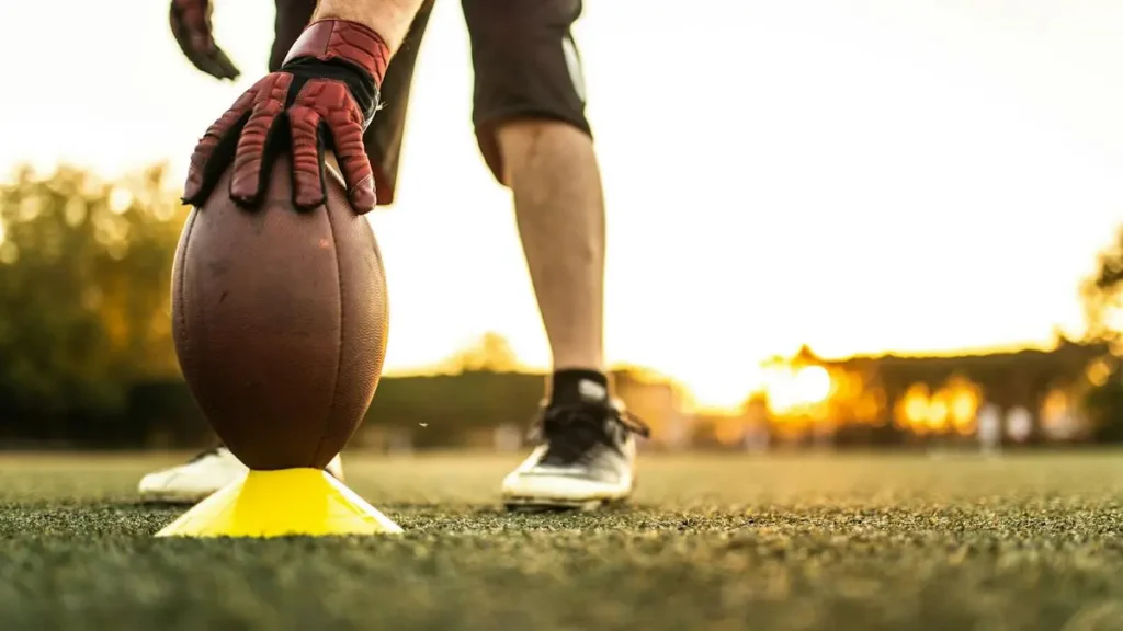 A football player prepares to kick the ball, with the ball resting on a tee, set against a backdrop of a sunset on a football field.