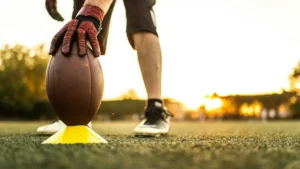 A football player prepares to kick the ball, with the ball resting on a tee, set against a backdrop of a sunset on a football field.