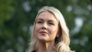The photo shows a person with blonde hair, wearing a small earring, against a blurred green background.