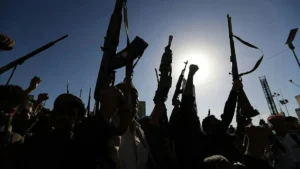 A group of people raise their weapons and fists in a demonstration under a bright sun.