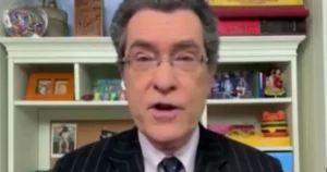 The image shows a person in a suit and tie, speaking in front of a bookshelf filled with various items and decorations.