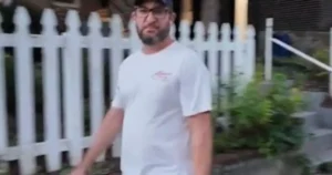 A man wearing a white t-shirt and a cap stands in front of a white picket fence, with a house and some greenery in the background.