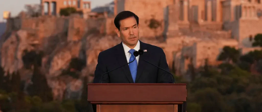 A man in a suit stands at a podium in front of a scenic backdrop, delivering a speech.