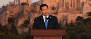 A man in a suit stands at a podium in front of a scenic backdrop, delivering a speech.