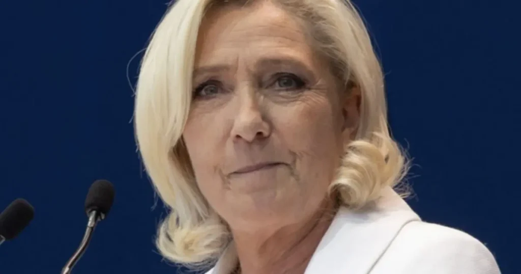 The image shows a person with blonde hair speaking at a podium with microphones.