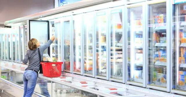 A woman shops in a grocery store's refrigerated section, reaching for an item from a glass-door freezer.