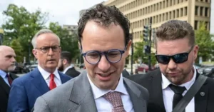 A man in a suit and glasses is walking outdoors, accompanied by security personnel, with a cityscape and trees in the background.