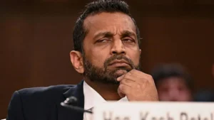 The image shows a man in a suit, appearing thoughtful, with his hand on his chin, seated at a table with a nameplate in front of him.