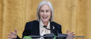 A woman in a professional setting is speaking at a podium with a microphone, gesturing with her hands.