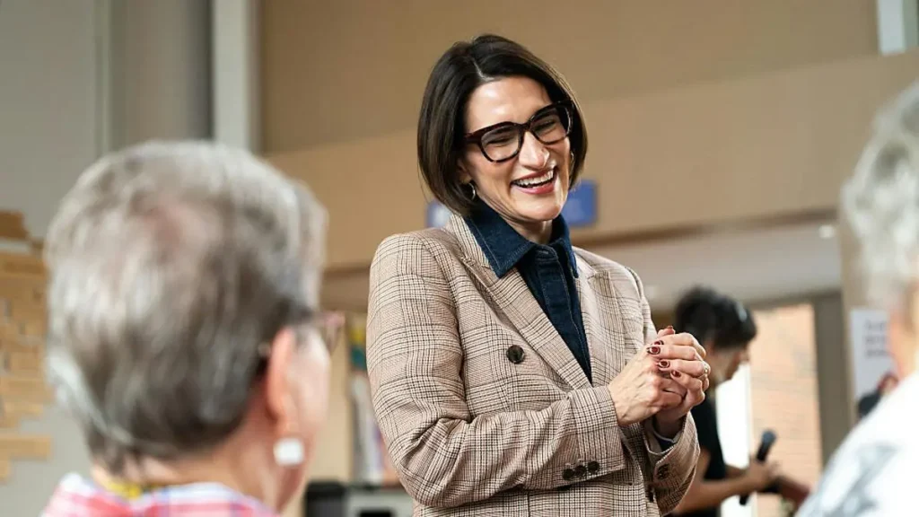 A woman in a plaid blazer smiles warmly while engaging in conversation with another person.