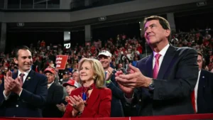 A group of people, including a man in a suit and a woman in a red jacket, are clapping at a political rally.