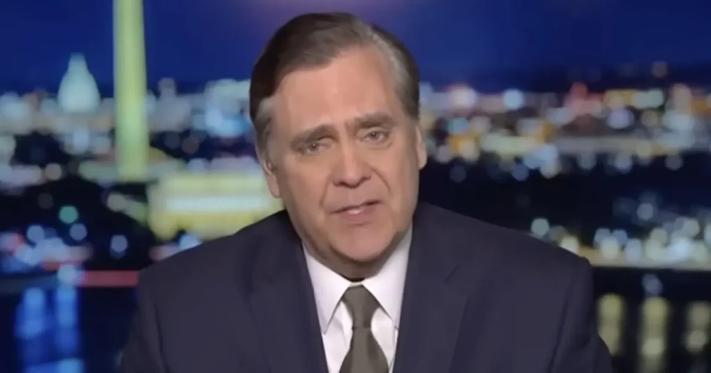 The image shows a man in a suit and tie, seated in front of a blurred cityscape background, likely during a news broadcast.