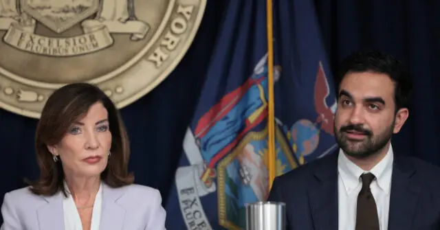 The photo shows two individuals seated at a press conference with the New York state seal and flag in the background.