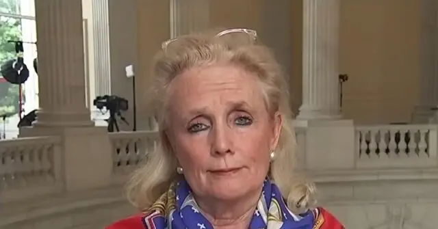 The image shows a person wearing a colorful scarf and pearl earrings, standing in front of a building with columns and a camera setup in the background.