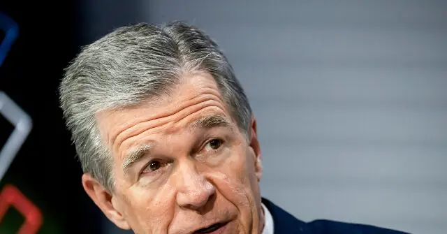 The image shows a close-up of a man with gray hair, wearing a suit, speaking at a press conference.